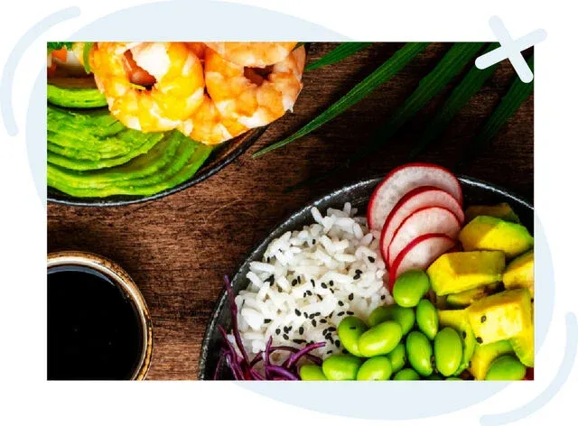 Colorful poke-style bowl with white rice topped with black sesame, edamame, radish slices, avocado chunks, purple cabbage, and shrimp, arranged on a rustic wooden table with a small dish of soy sauce.