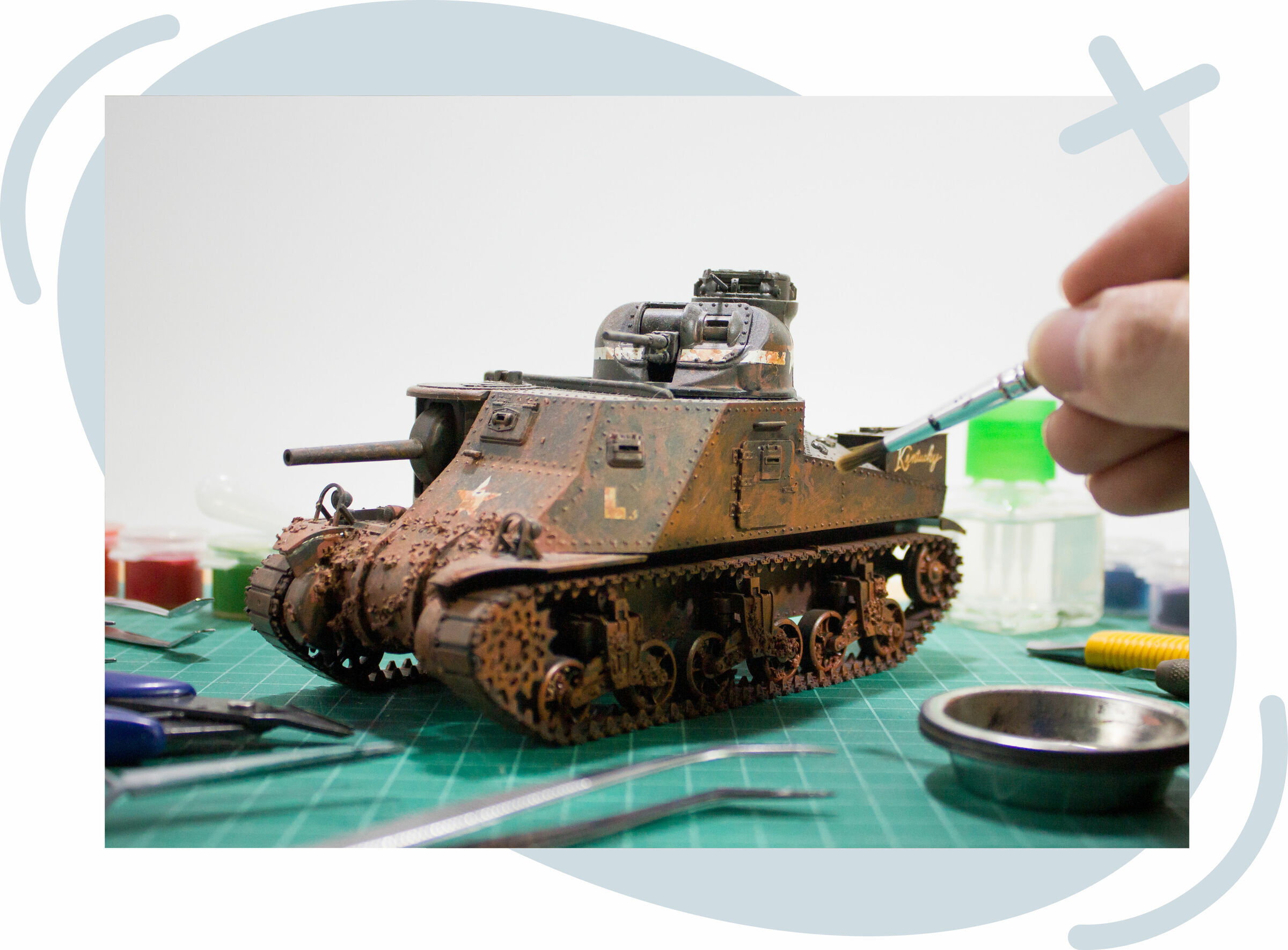 Close-up of a hand painting a weathered scale model tank on a green cutting mat, surrounded by hobby tools and paints.