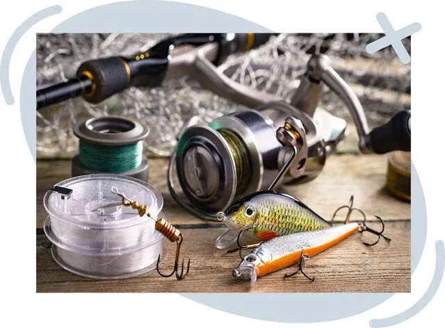 Close-up of fishing tackle on a wooden table, including a spinning reel, fishing line spools, and colorful hard bait lures with treble hooks, set against a tangle of netting in the background.