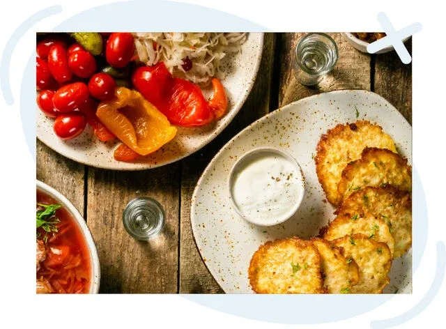 Rustic table spread with Eastern European dishes including potato pancakes with sour cream, pickled vegetables, and small glasses of clear liquor.