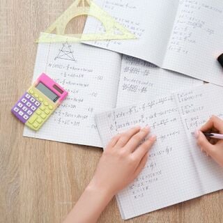 Hands working on algebra and geometry problems in grid notebooks on a wooden desk with a calculator, highlighters, and triangular protractor.