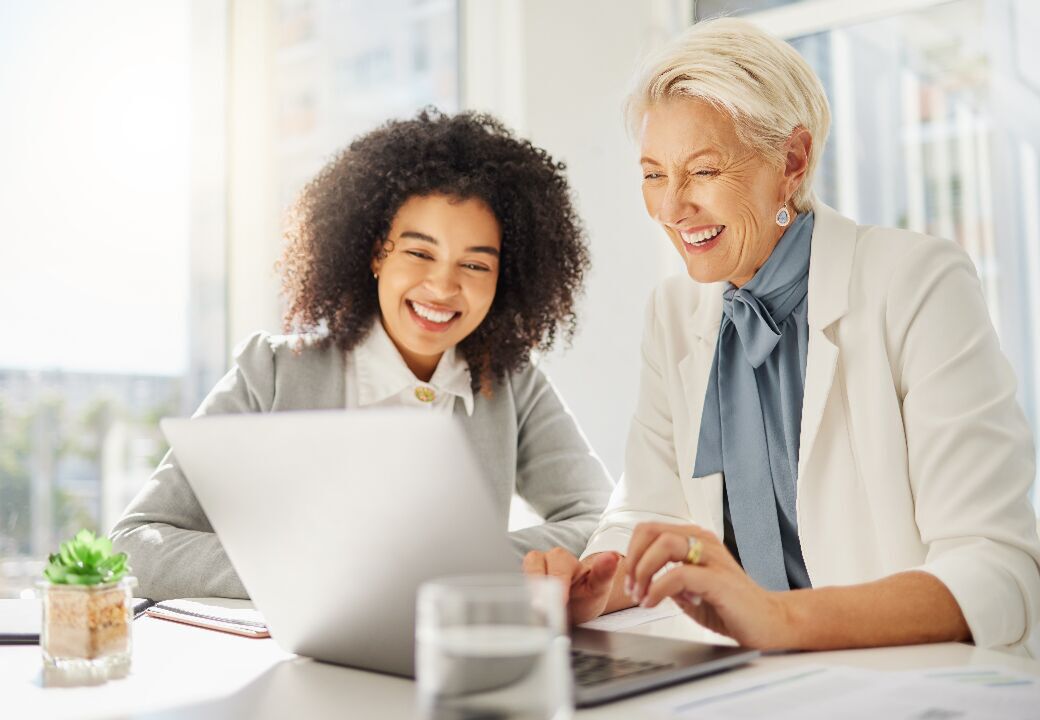 Two colleagues smiling and working together on a laptop in a bright office.
