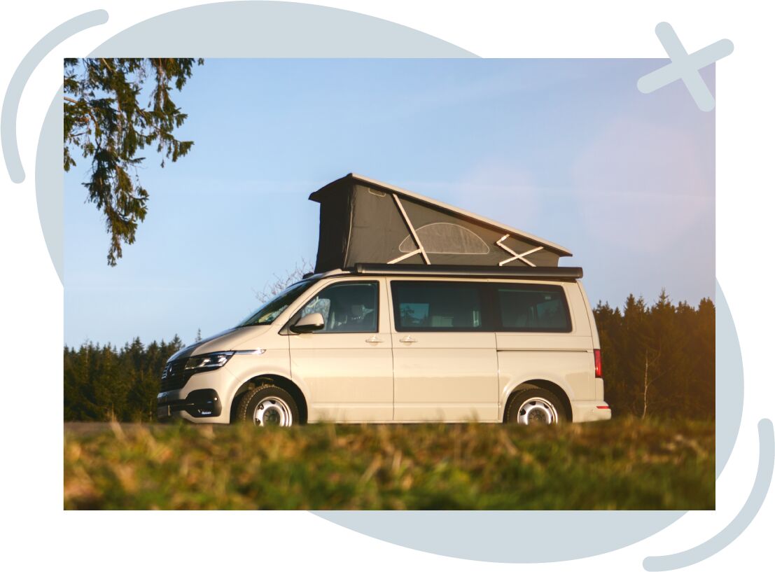 Beige camper van with a raised pop-up roof parked on grass in front of a forest under a clear blue sky at sunset.