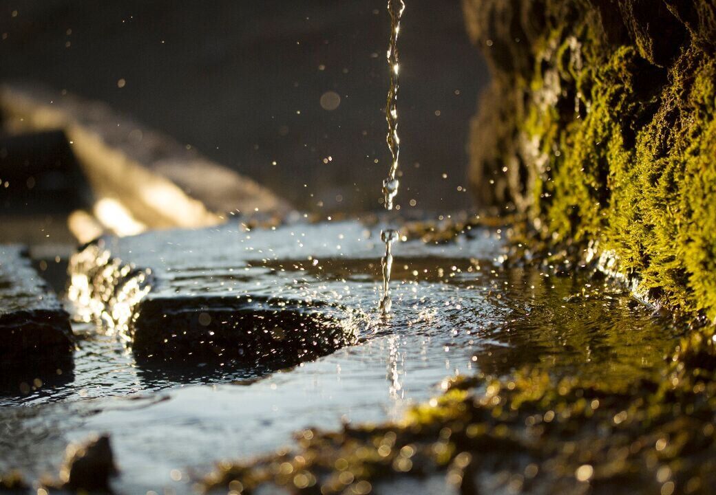 Close-up of water droplets falling into a shallow stream beside moss-covered rock in warm sunlight