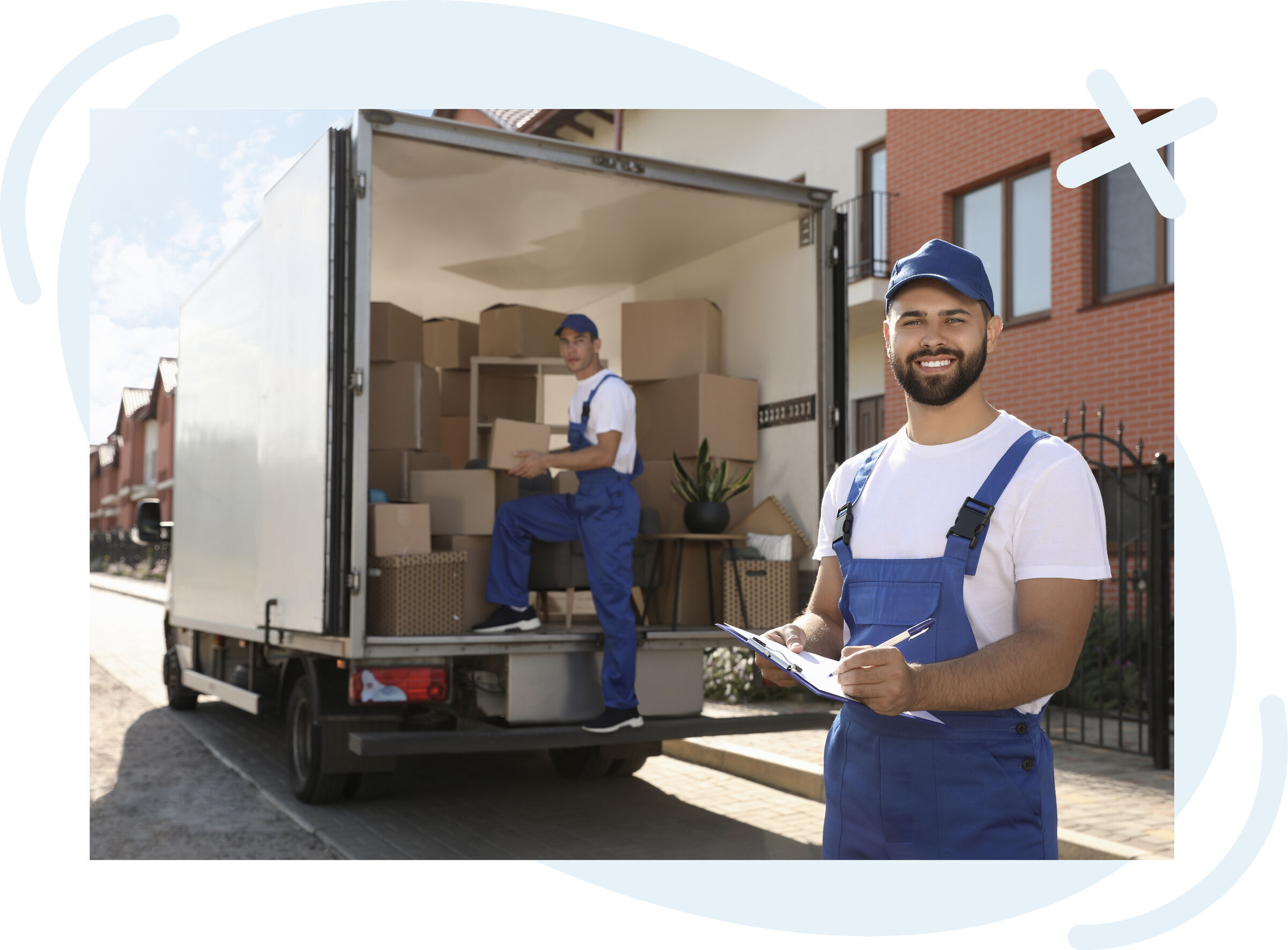 Two movers in blue uniforms working by a loaded moving truck on a residential street; one smiles at the camera holding a clipboard while the other arranges boxes inside the truck.