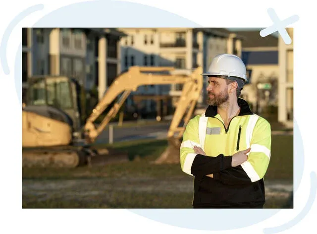 Construction worker in high-visibility jacket and white hard hat standing with arms crossed at a job site with excavators in the background.
