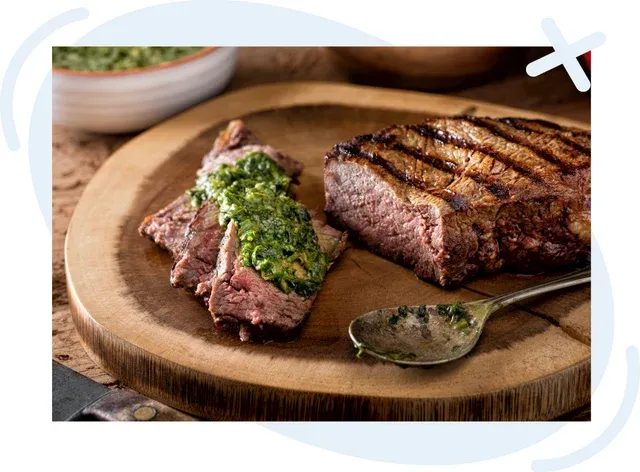 Sliced grilled steak topped with green chimichurri sauce on a wooden board, with a spoon and more sauce in the background.