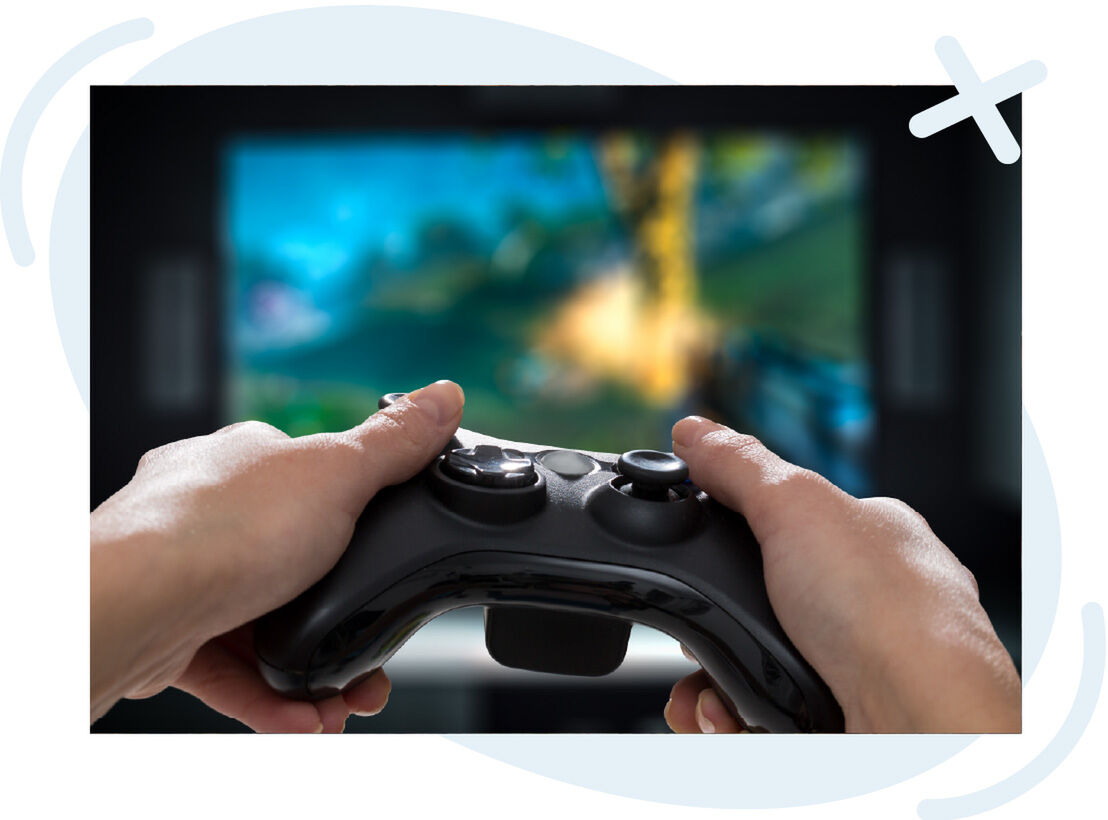 Close-up of hands holding a black game controller in front of a blurry TV screen showing a colorful game scene.