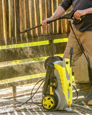 Person pressure-washing a wooden gate outdoors using a yellow pressure washer.