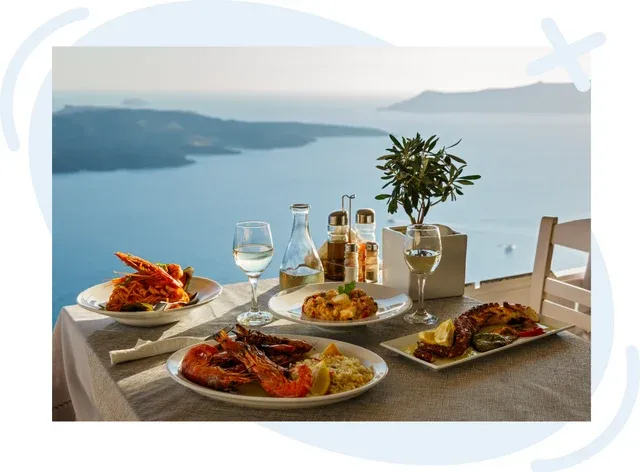 Seaside table set with Mediterranean seafood dishes, wine glasses, and condiments overlooking a calm blue sea and distant islands at sunset.