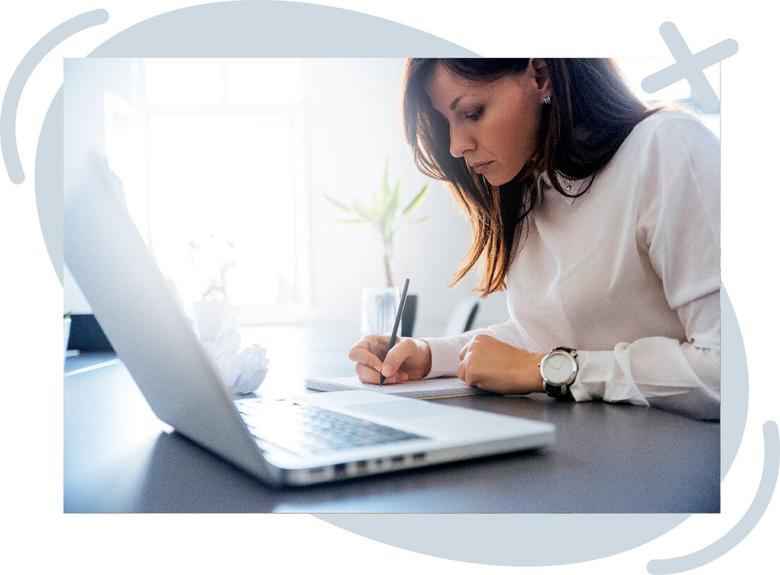Person writing in a notebook beside an open laptop on a bright desk.