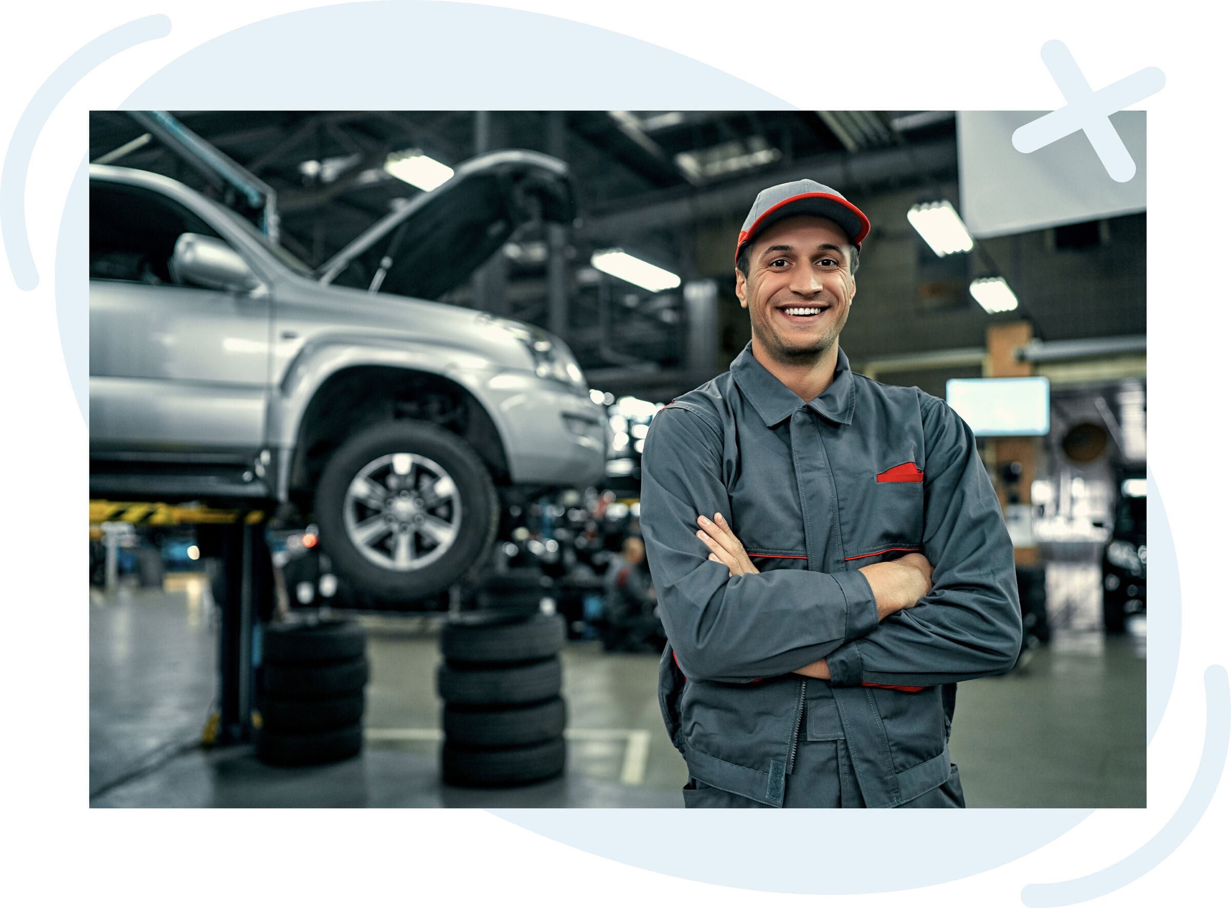 Smiling auto mechanic in a gray uniform and cap stands with arms crossed in a service garage, with a silver SUV lifted for repair in the background.