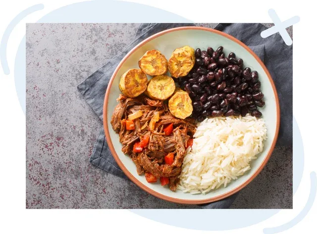 Plate with Venezuelan-style meal: shredded beef with peppers and onions, white rice, black beans, and fried plantain slices, shot from above on a gray surface.