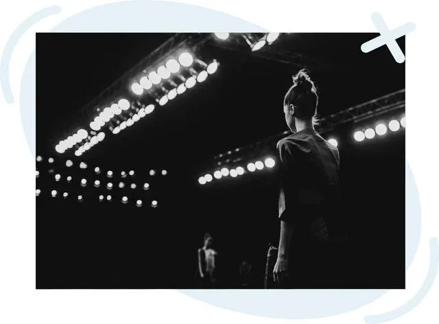 Black-and-white photo of a fashion show runway with bright spotlights and a model standing near the stage edge, seen from behind.