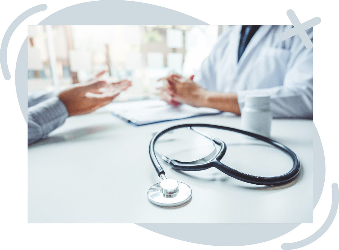 Close-up of a stethoscope on a clinic desk while a patient and a healthcare professional discuss something in the background. The doctor wears a white coat, and a pill bottle and clipboard are on the table. The scene is softly lit and slightly out of focu
