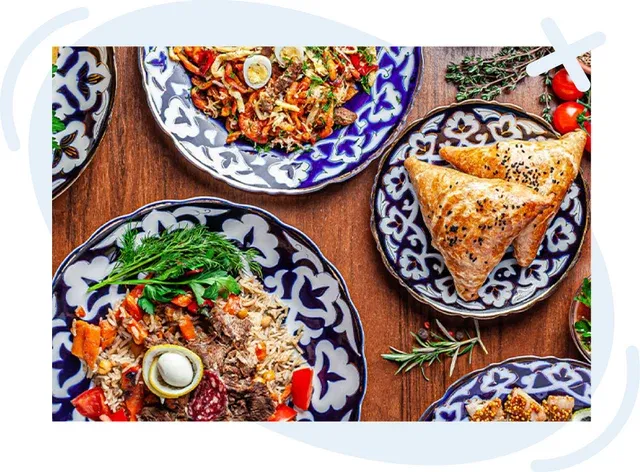 Assorted Central Asian dishes on patterned blue-and-white plates arranged on a wooden table, including rice pilaf with meat, baked triangular pastries, and colorful vegetable stir-fries with herbs and garnishes.