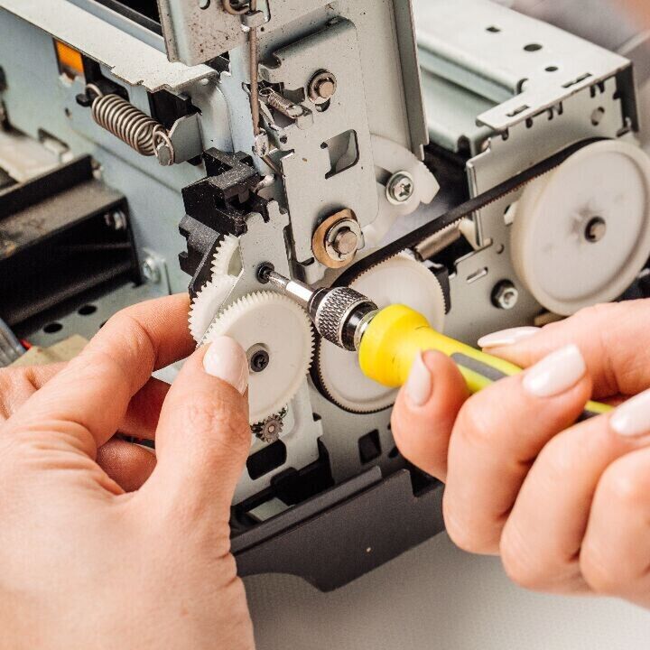 Close-up of hands repairing the internal gears of a mechanical device with a yellow-handled screwdriver.