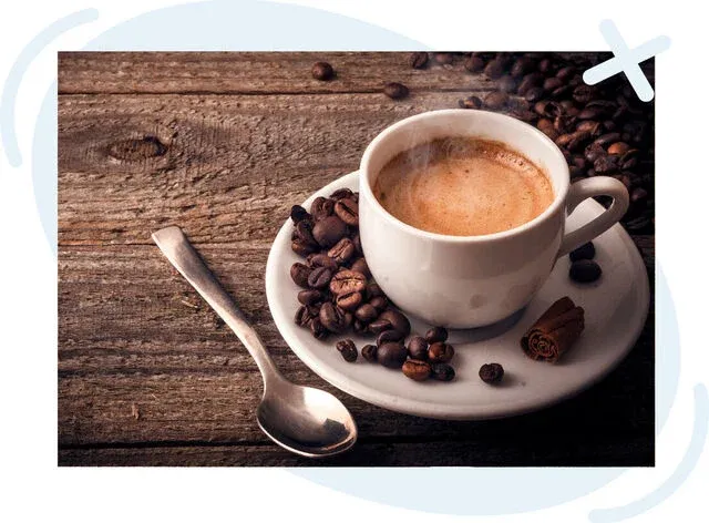 A steaming espresso in a white cup and saucer on rustic wooden table, surrounded by roasted coffee beans, a spoon, and a cinnamon stick.