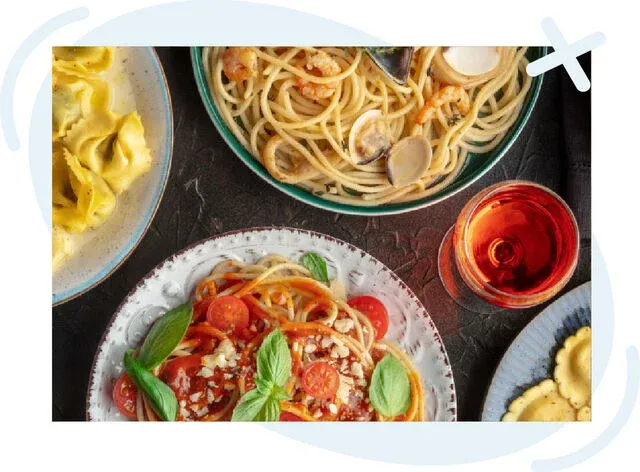 Top-down view of an Italian meal spread with various pasta dishes and a glass of red beverage on a dark table.