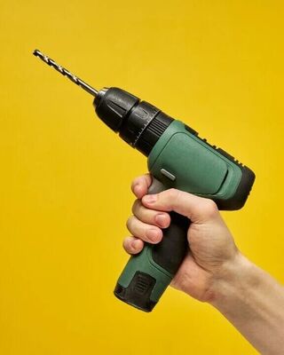 Hand holding a green cordless power drill with a metal drill bit against a solid yellow background.