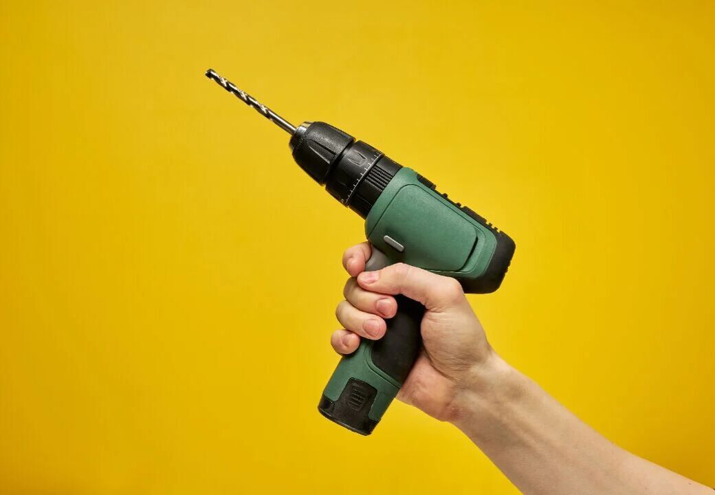 Hand holding a green cordless power drill with a metal drill bit against a solid yellow background.