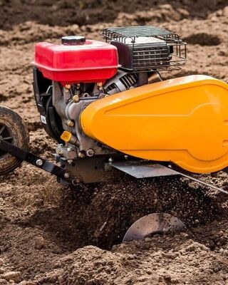Close-up of a motorized garden tiller churning soil in a field, with a red fuel tank and bright yellow engine cover.