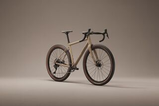 Desert tan gravel bike with drop bars and chunky tires posed three-quarter view on a smooth gray studio backdrop.
