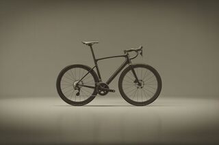 Side profile of a carbon black road bicycle on a smooth mid-grey studio backdrop, fully visible wheels and drop handlebars lit by soft, even lighting.