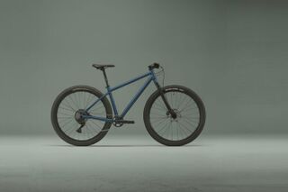Side view of a matte blue hardtail mountain bike in a minimalist studio setting.