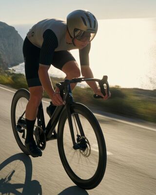 Cyclist in aerodynamic gear riding a road bike along a coastal highway at sunrise, ocean shimmering in the background.