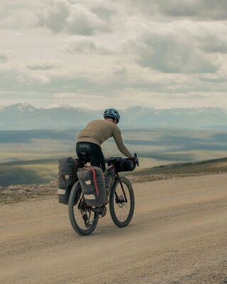 Bikepacker riding a loaded gravel bike along a wide, dusty mountain road with expansive rolling hills and snow-capped peaks under a cloudy sky.