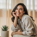 Smiling person in a relaxed indoor setting, leaning on their hand beside a small potted succulent.