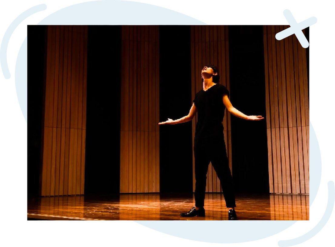 Solo performer on a wooden stage, arms outstretched, looking upward under warm spotlight.