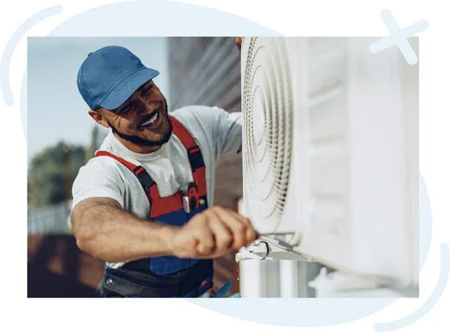 HVAC technician smiling while using a screwdriver to service an outdoor air conditioning unit mounted on a house exterior.