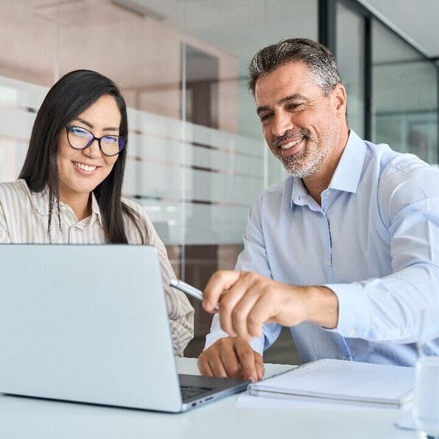 Two colleagues smiling while reviewing something on a laptop in a modern glass-walled office.