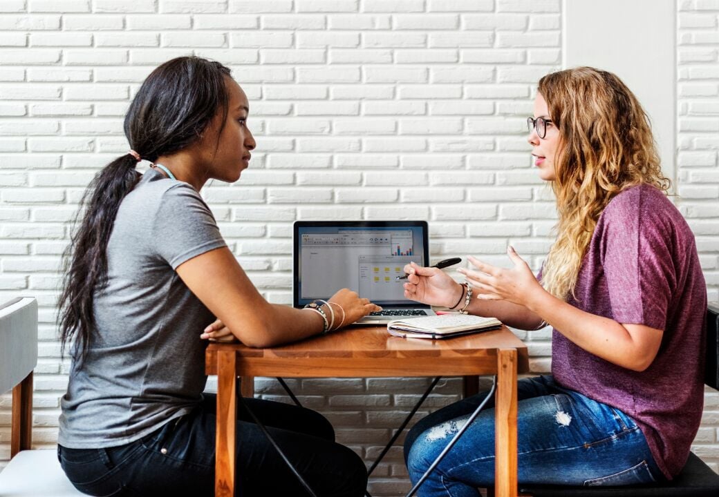 Two people sitting at a small table discussing work with an open laptop between them.