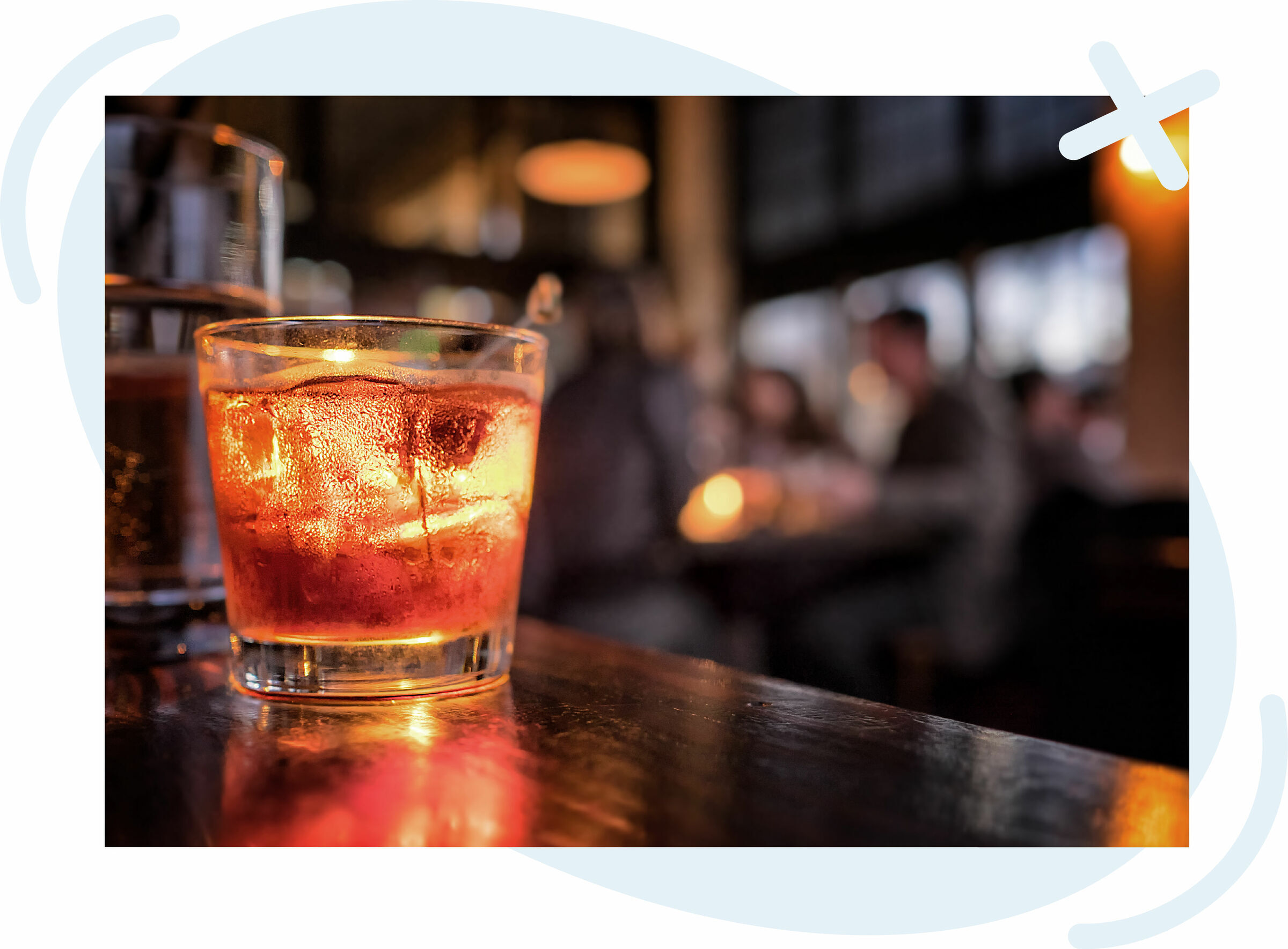 Close-up of a cocktail with ice in a low glass on a bar, glowing amber under warm lights, with a blurred, cozy bar scene and patrons in the background.