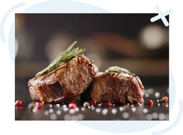 Two seared steak medallions garnished with rosemary on a dark surface, sprinkled with coarse salt and pink peppercorns.