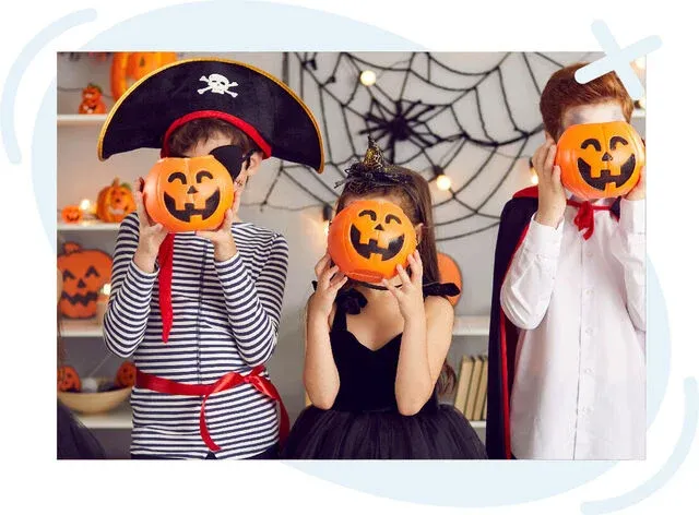 Three children in Halloween costumes hold jack-o'-lantern props in front of their faces, standing in a festively decorated room with spiderwebs and pumpkins.
