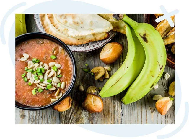 Top-down view of a rustic table spread with African-inspired dishes, including a bowl of tomato-peanut stew, green plantains, fried dough bites, and a folded flatbread.