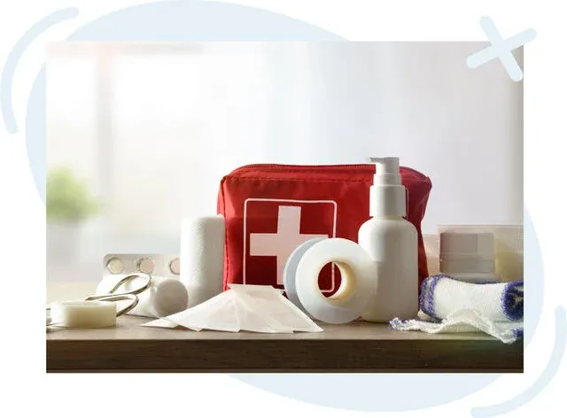 First aid kit on a wooden surface with medical supplies including bandages, adhesive tape, scissors, gauze, blister-pack tablets, and a pump bottle, with a red pouch marked with a white cross in the background.