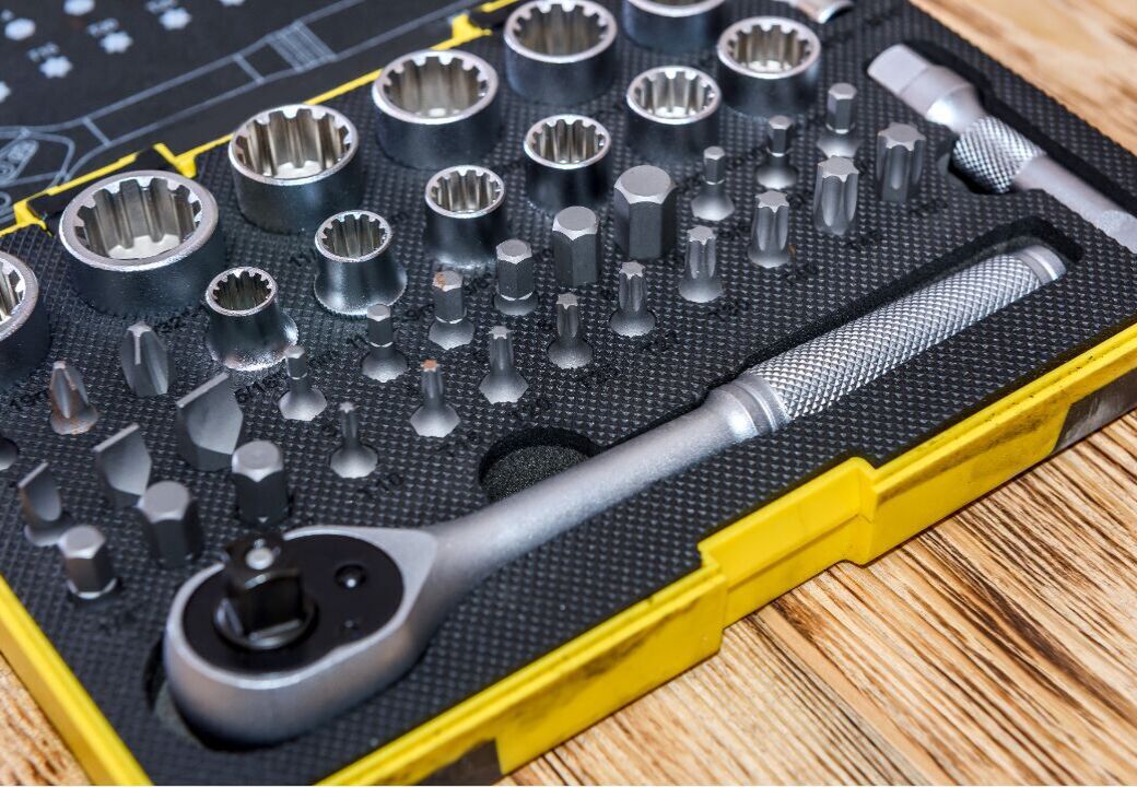 Close-up of a socket wrench set in a yellow plastic case, showing a ratchet handle and various sockets and bits organized in foam on a wooden surface.
