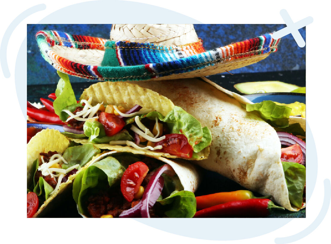Close-up of vibrant Mexican food: hard-shell tacos and a burrito stuffed with lettuce, tomatoes, onions, cheese, and meat, garnished with colorful chili peppers and topped with a woven sombrero.