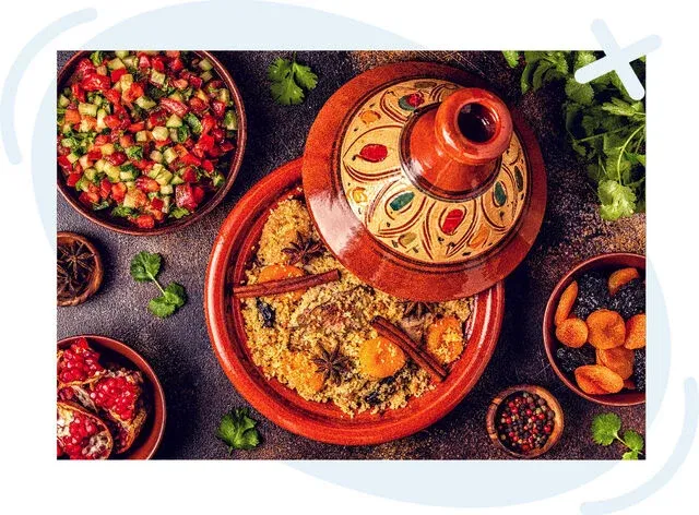 Top-down view of a Moroccan tagine meal with couscous and spices in a decorated clay pot, surrounded by salads, dried fruit, and herbs.