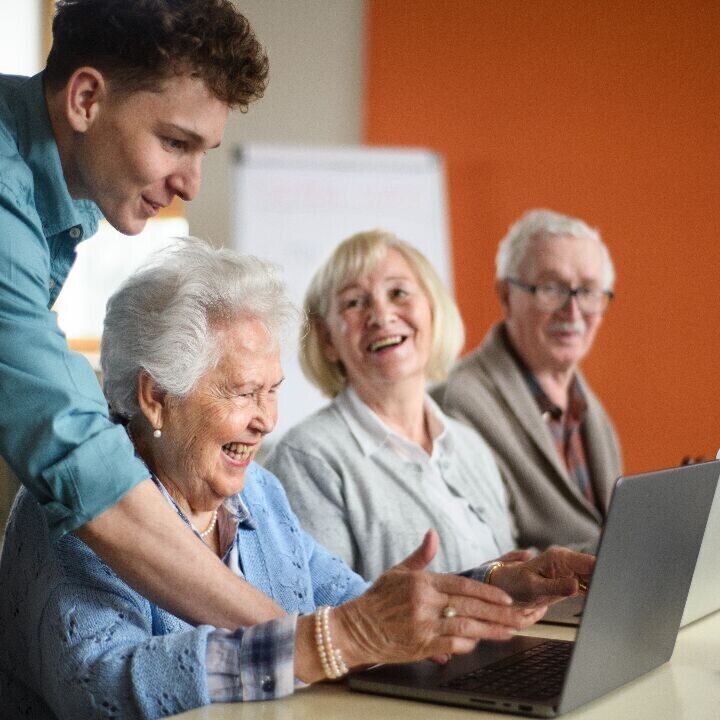 Young man helping a group of seniors using laptops in a cheerful classroom setting.