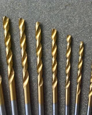 Set of gold-colored twist drill bits arranged in a row from largest to smallest on a gray textured surface.