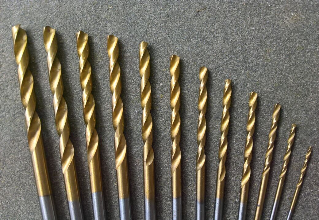 Set of gold-colored twist drill bits arranged in a row from largest to smallest on a gray textured surface.