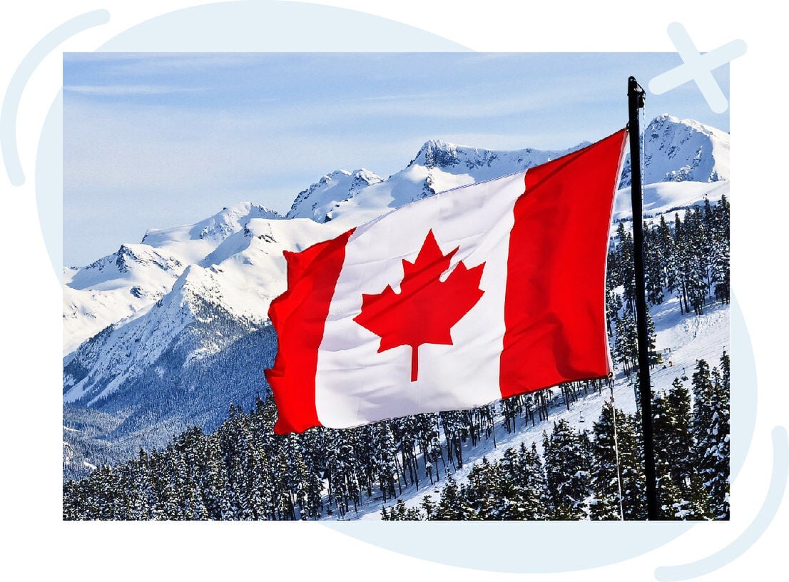 Canadian flag waving in front of snow-covered mountains and pine forests on a clear winter day.