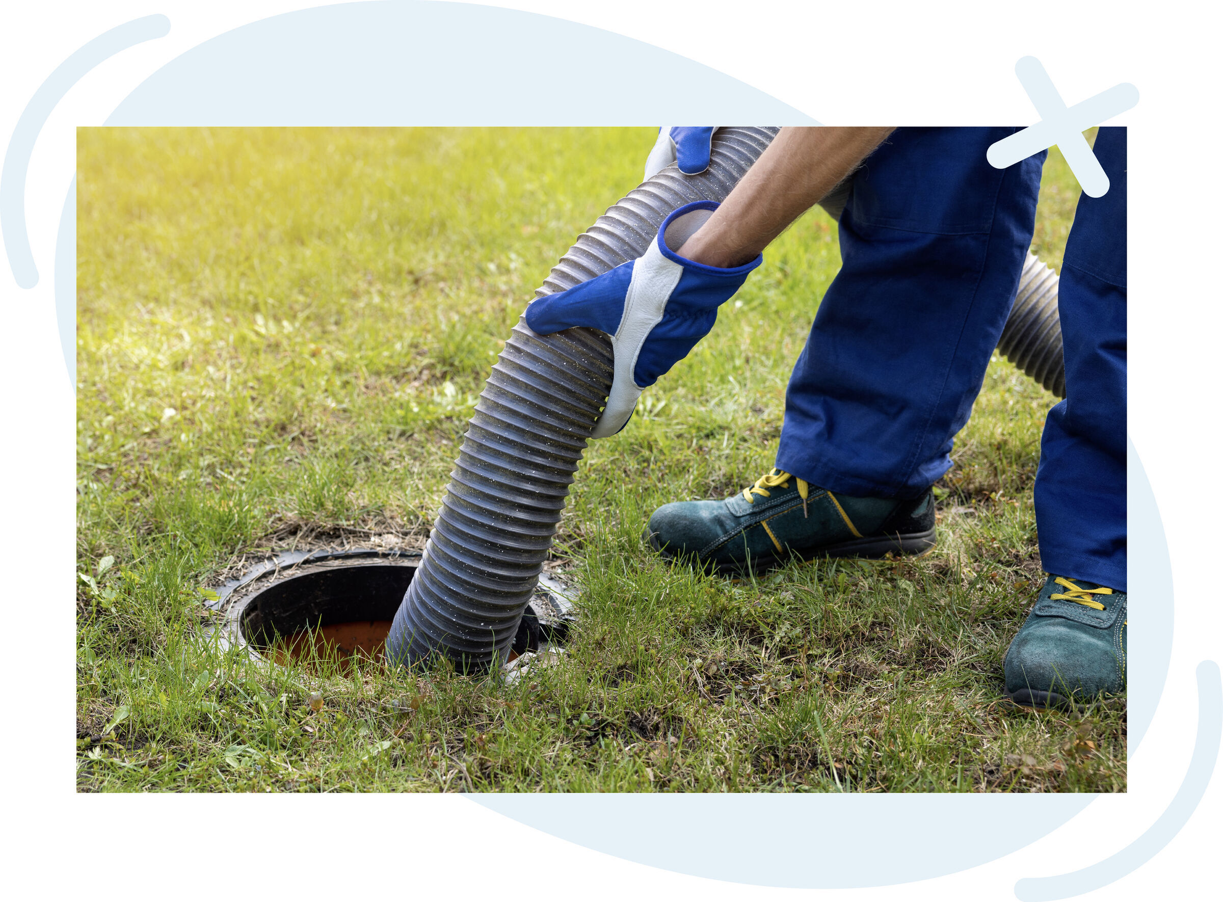 Worker in blue coveralls and gloves inserting a large corrugated hose into an outdoor septic tank opening on a grassy lawn.