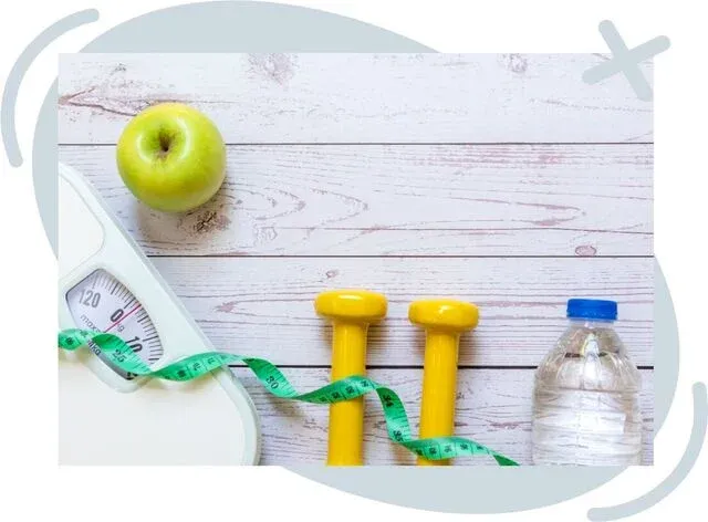 Flat lay of healthy lifestyle items on a light wooden surface: a green apple, a bathroom scale partly visible with a green measuring tape draped across it, two small yellow dumbbells, and a plastic water bottle with a blue cap. Soft gray abstract shapes f